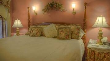 Een slaapkamer met een groot bed, kussens met een bloemenprint, een nachtkastje met een lamp en wandlampen.