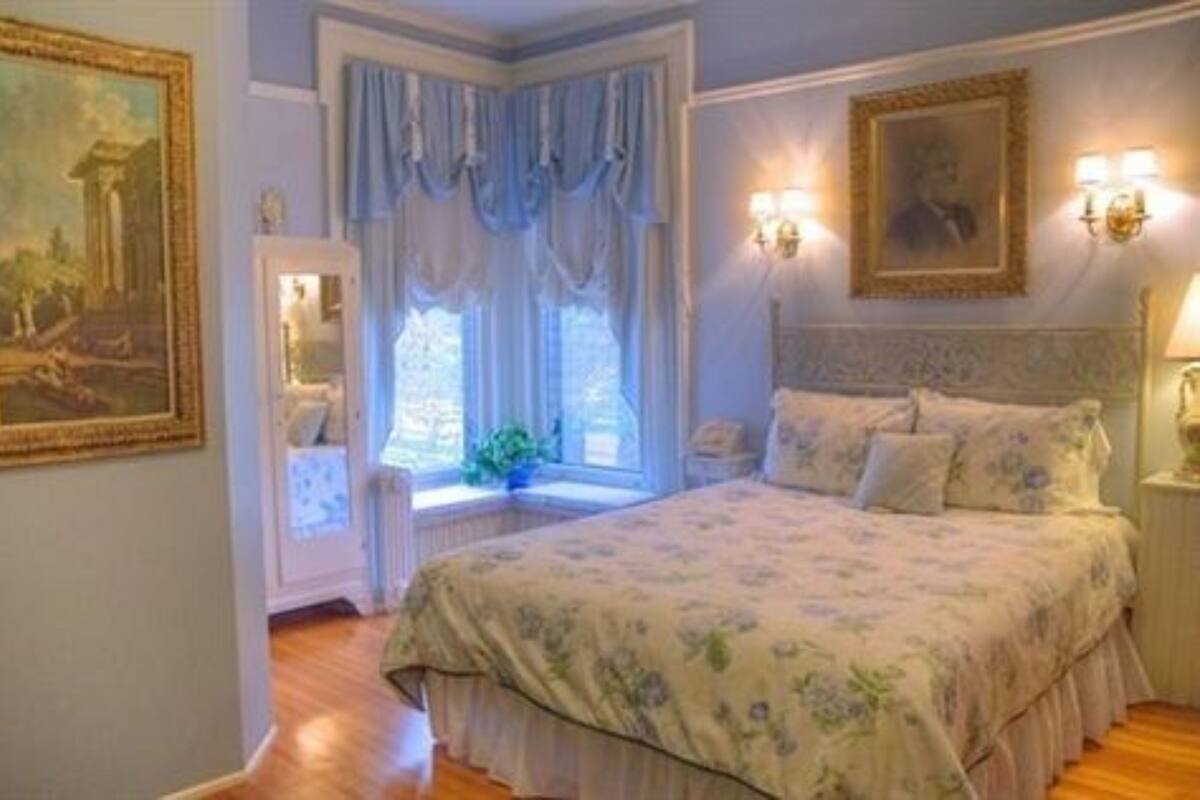 A bedroom with a large bed, a window with curtains, a painting on the wall, and a nightstand with a lamp.
