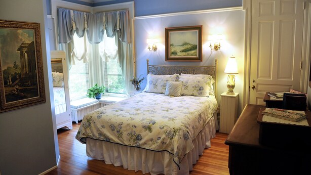 A bedroom with a large bed, a window with curtains, a painting on the wall, and a nightstand with a lamp.