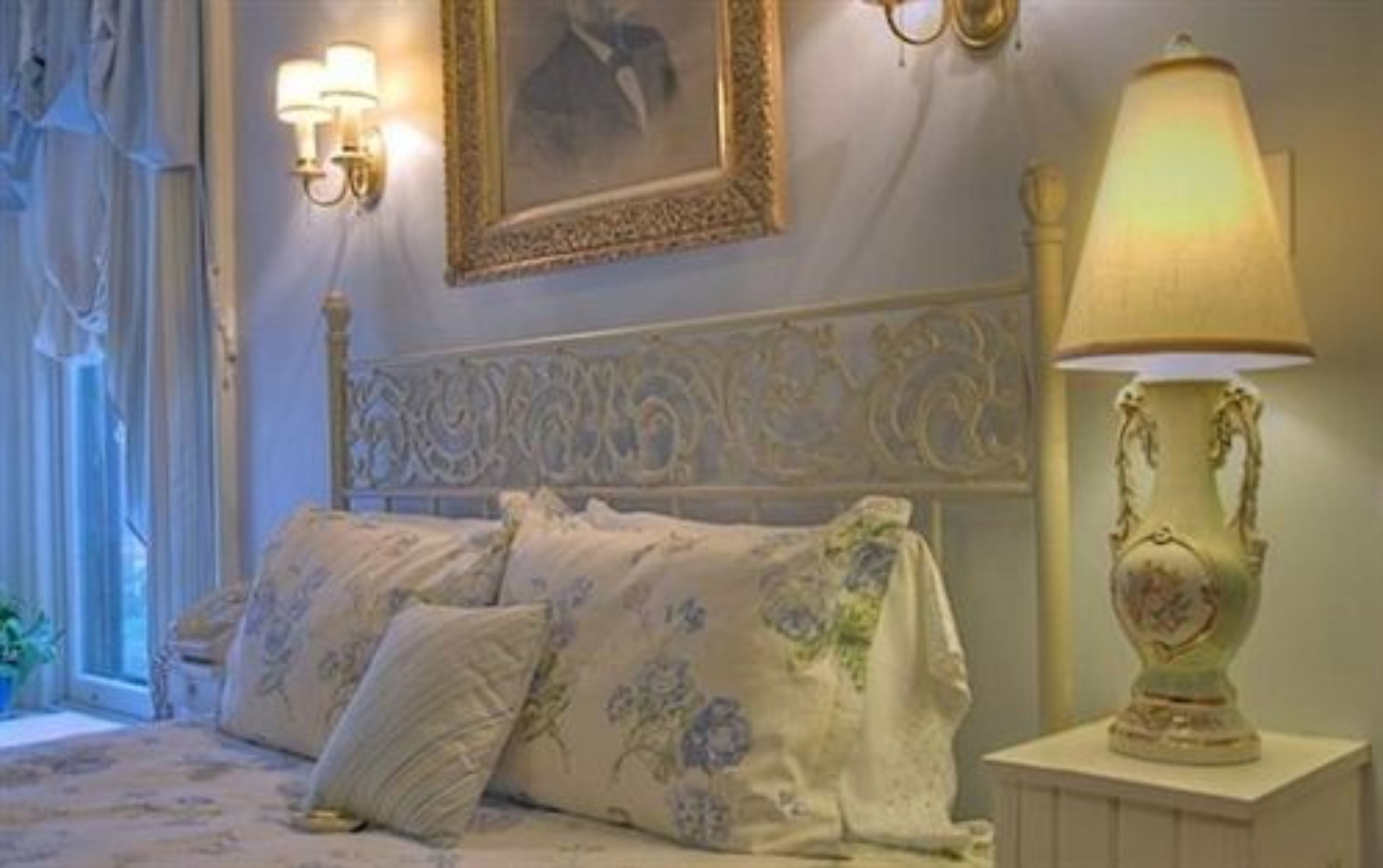 Un dormitorio con una cama, ropa de cama estampada con motivos florales, una lámpara decorativa y un cuadro en la pared.