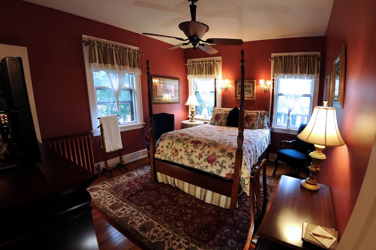 A four-poster bed with floral bedding, a ceiling fan, and a nightstand with a lamp.