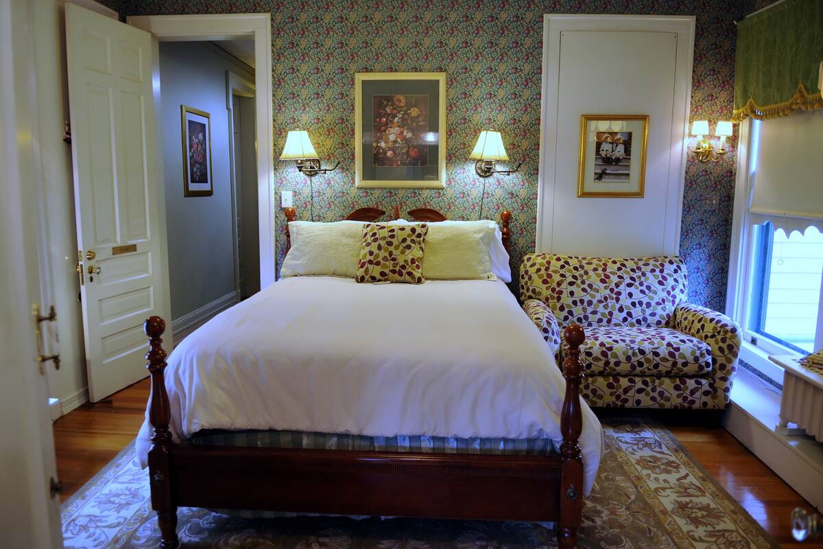A bedroom with a four-poster bed, two wall-mounted lamps, a floral-patterned armchair, and a framed picture on the wall.