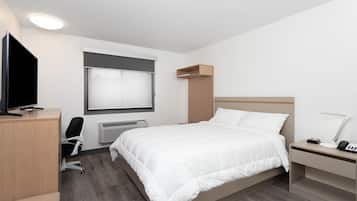 A modern hotel room with a bed, a desk, a chair, a TV, a bedside table with a lamp, and a window with blinds.