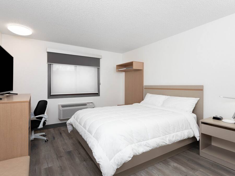 A modern hotel room with a bed, a desk, a chair, a TV, a bedside table with a lamp, and a window with blinds.
