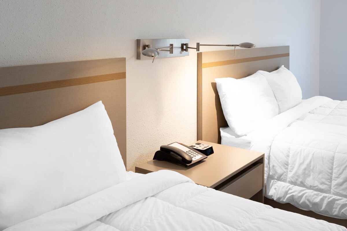 Camera d'albergo con due letti, un comodino con telefono e lampada, e un lampadario a parete.
