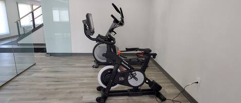 Two stationary bikes in a room with a wooden floor and a staircase in the background.