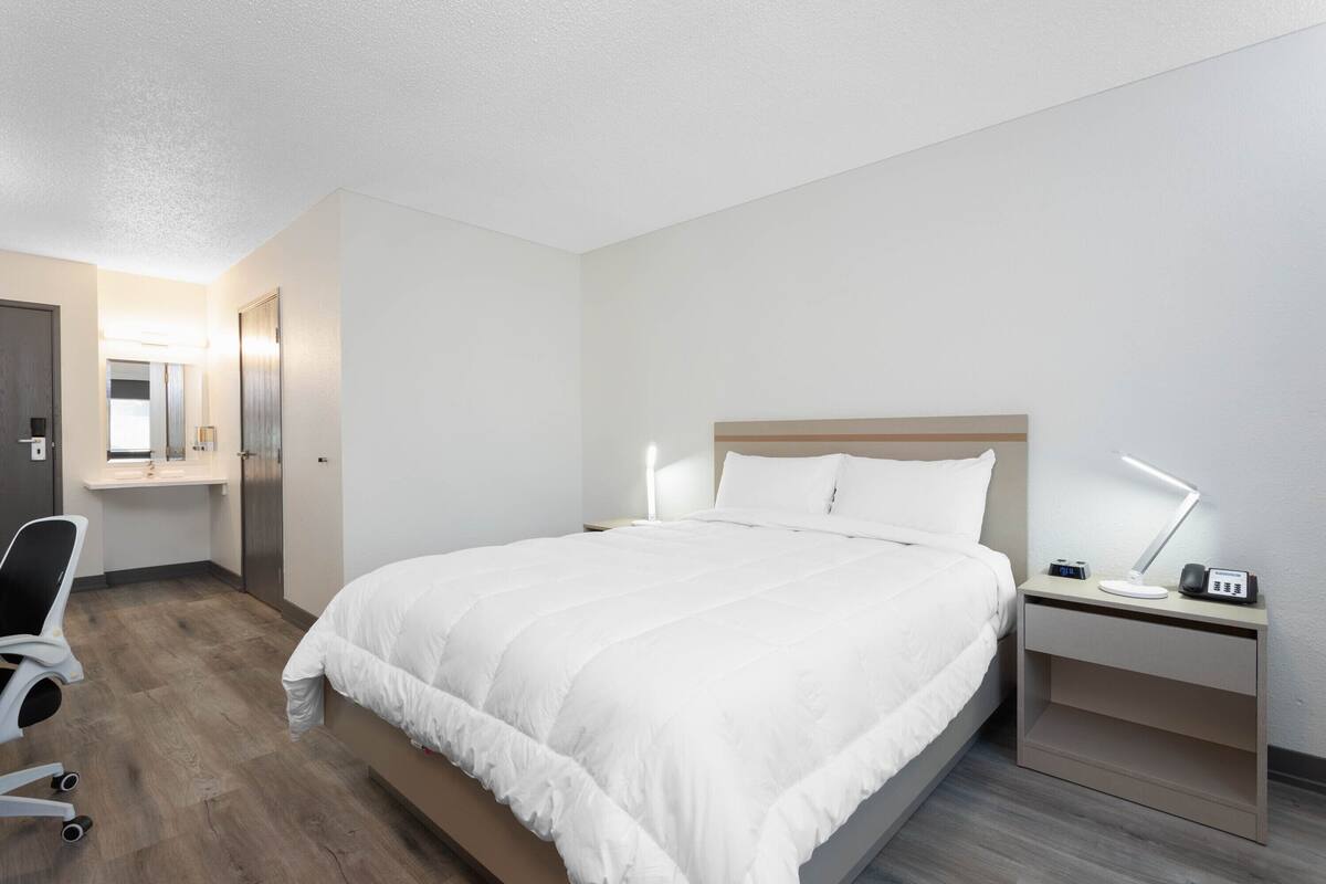 Habitación de hotel con cama, mesita de noche, lámpara, silla y escritorio con computadora.