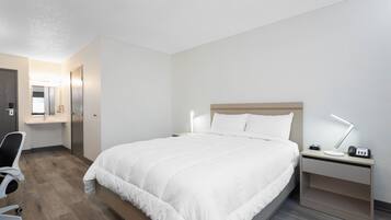 Habitación de hotel con cama, mesita de noche, lámpara, silla y escritorio con computadora.