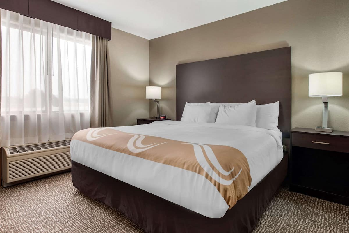 A hotel room with a large bed, a bedside table with a lamp, a heater, and a window with sheer curtains.