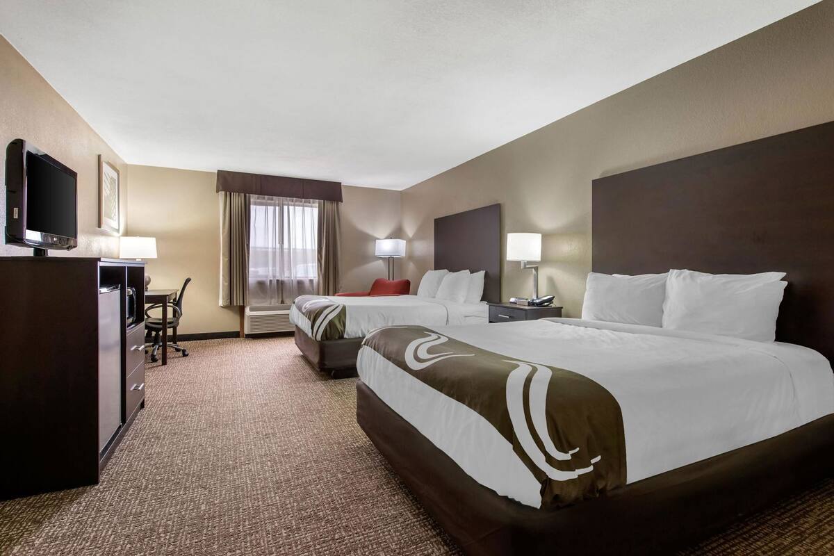 A hotel room with two beds, a desk, a TV, and a window with curtains.