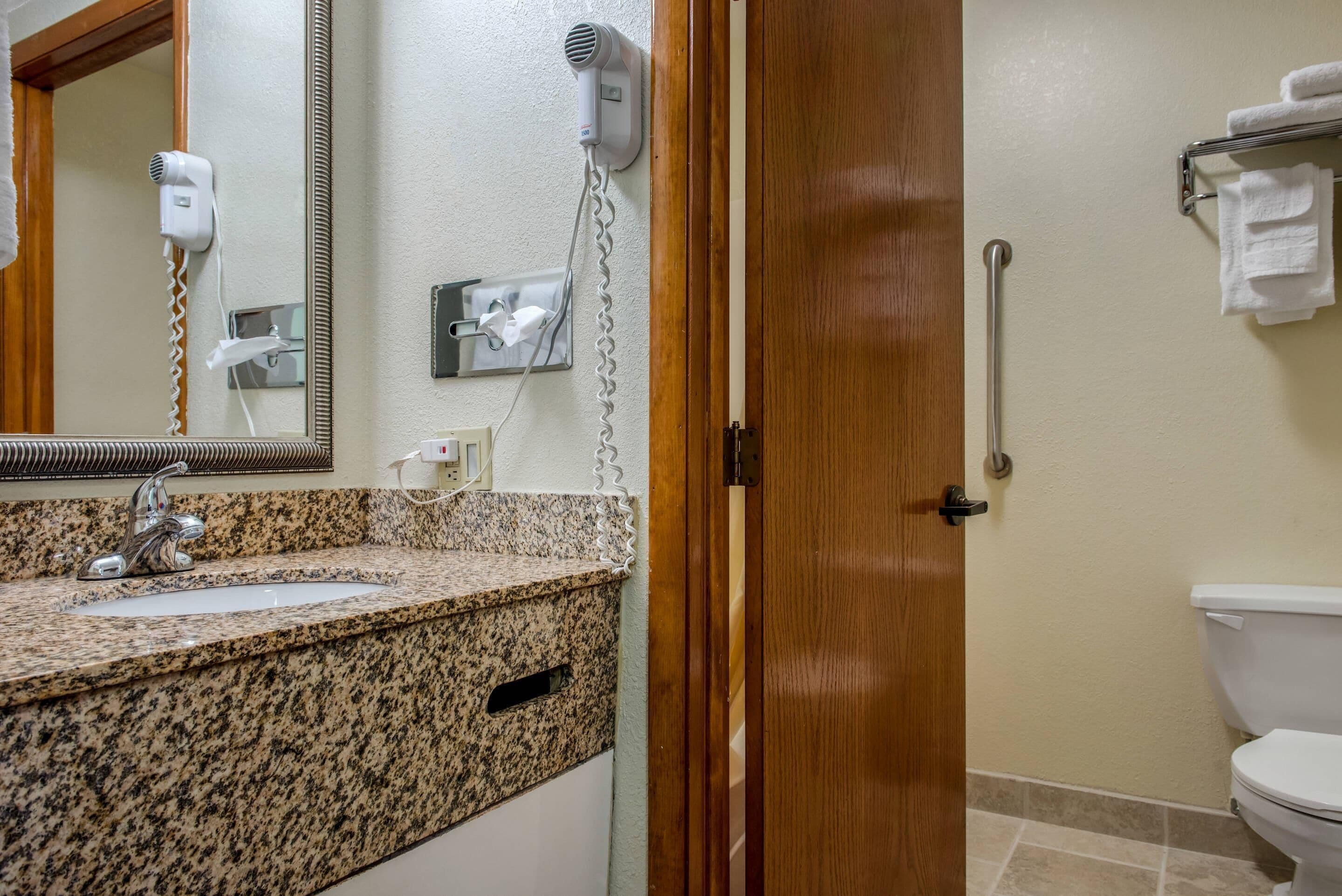 Een badkamer met een granieten aanrecht, een wastafel, een spiegel, een haardroger, een handdoekhouder en een handgreep.