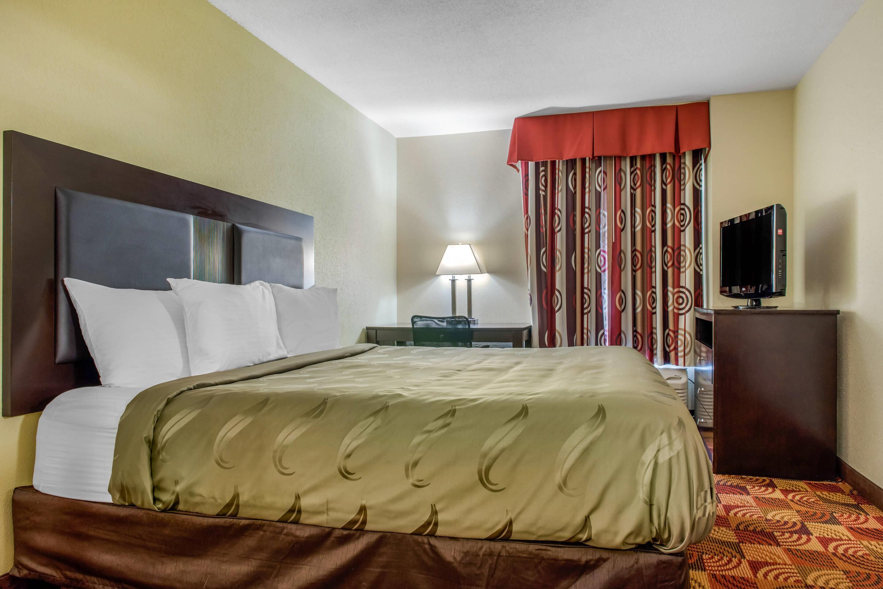 A hotel room with a bed, a desk, a lamp, a TV, and a patterned curtain.
