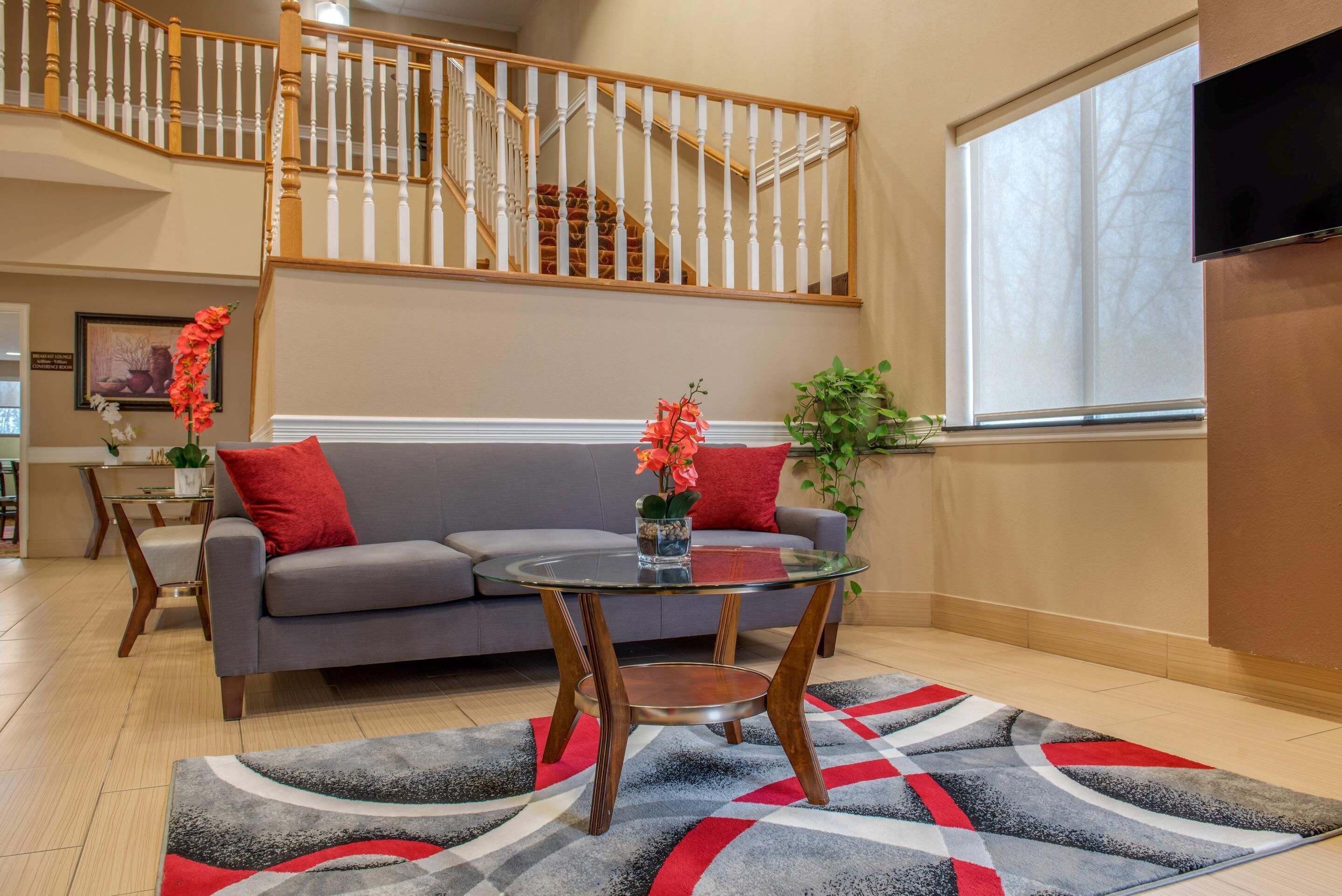 A living room with a gray sofa, a glass coffee table, and a red and gray rug. A staircase with wooden railings is visible in the background.