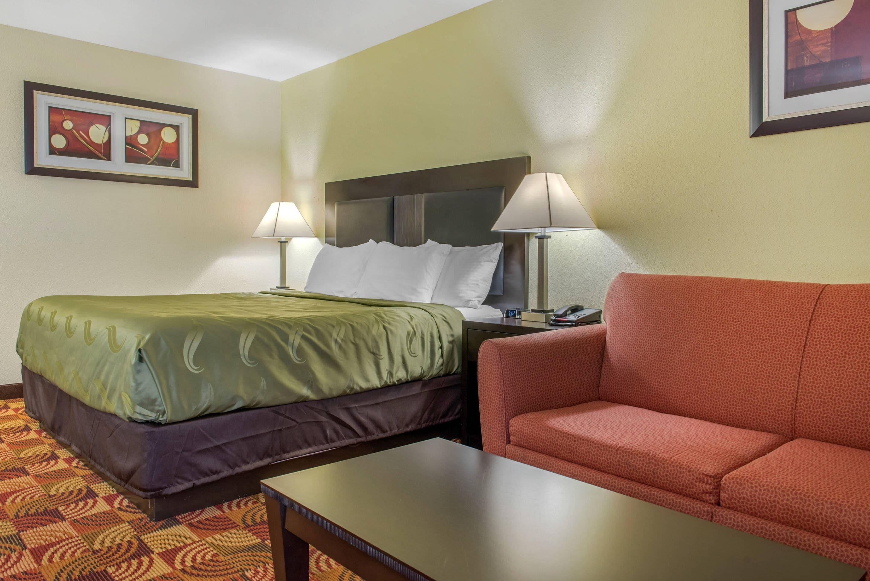 Une chambre d’hôtel avec un lit, deux lampes de chevet, un canapé rouge, une petite table et deux tableaux encadrés au mur.