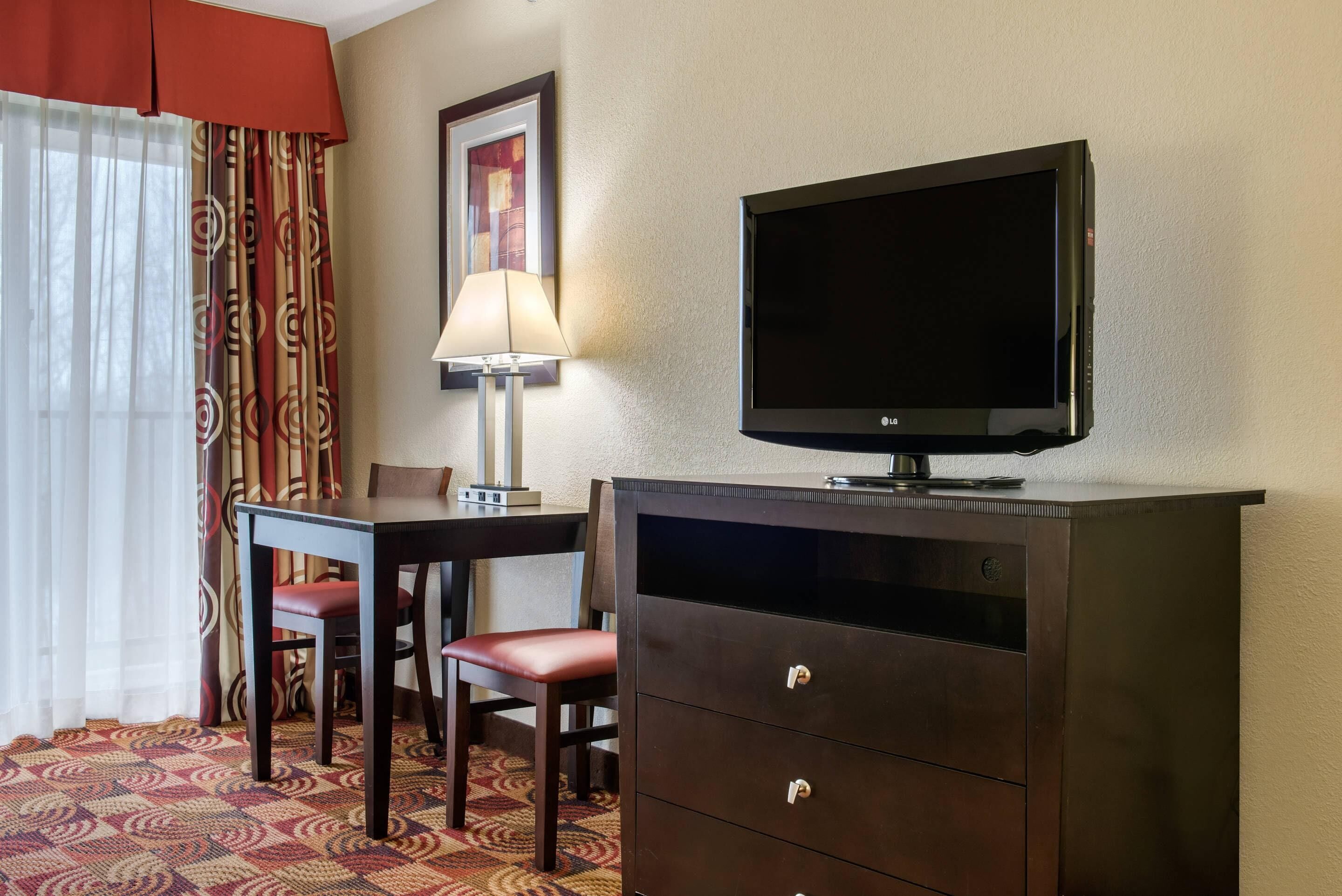 Habitación de hotel con televisión en un mueble, escritorio con silla, lámpara y alfombra estampada.