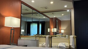 Habitación de hotel con una cama grande, una mesita de noche, un espejo, un baño con bañera y un ventanal con cortinas.