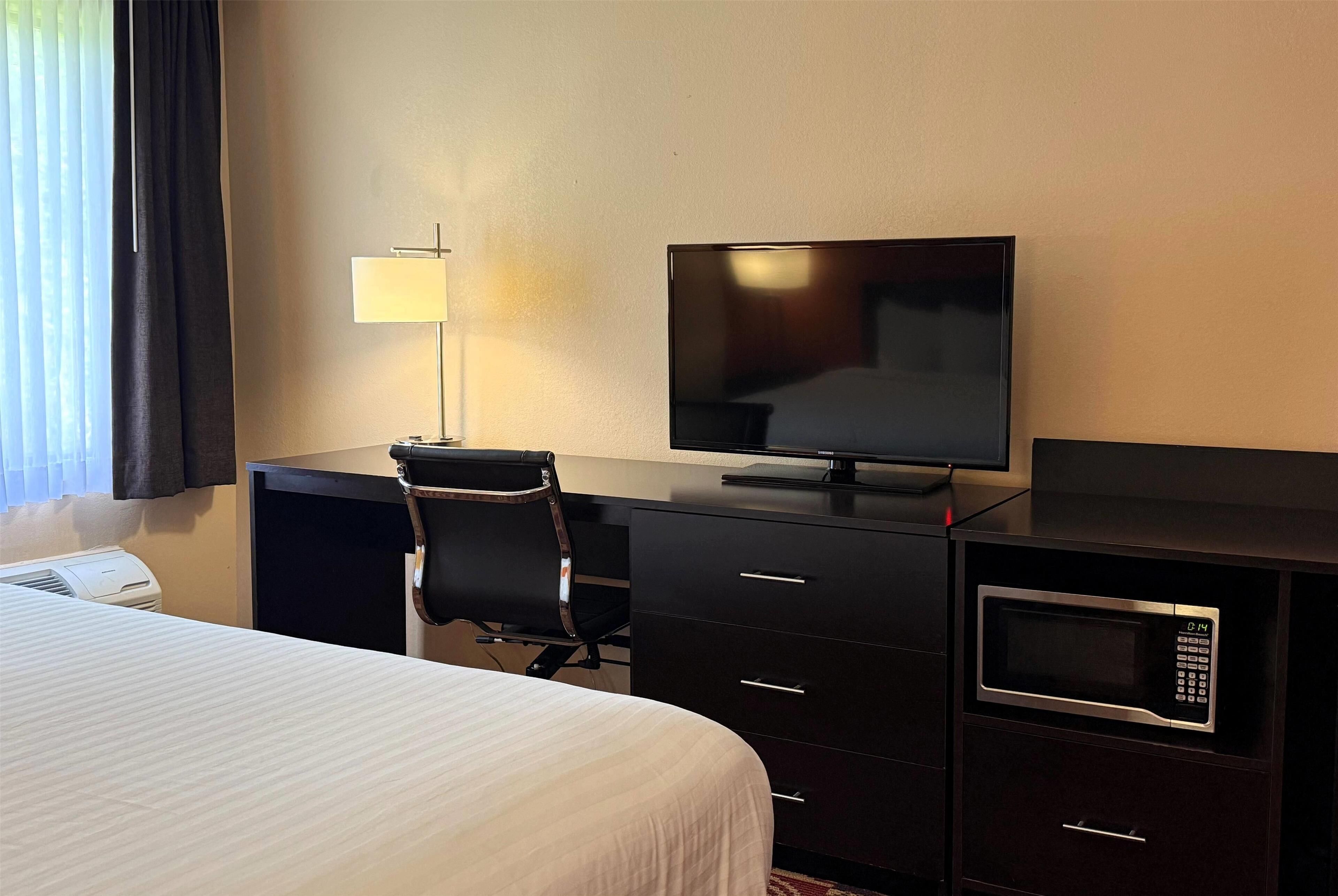 A hotel room with a bed, a desk with a chair, a television, a lamp, a microwave, and a small safe.