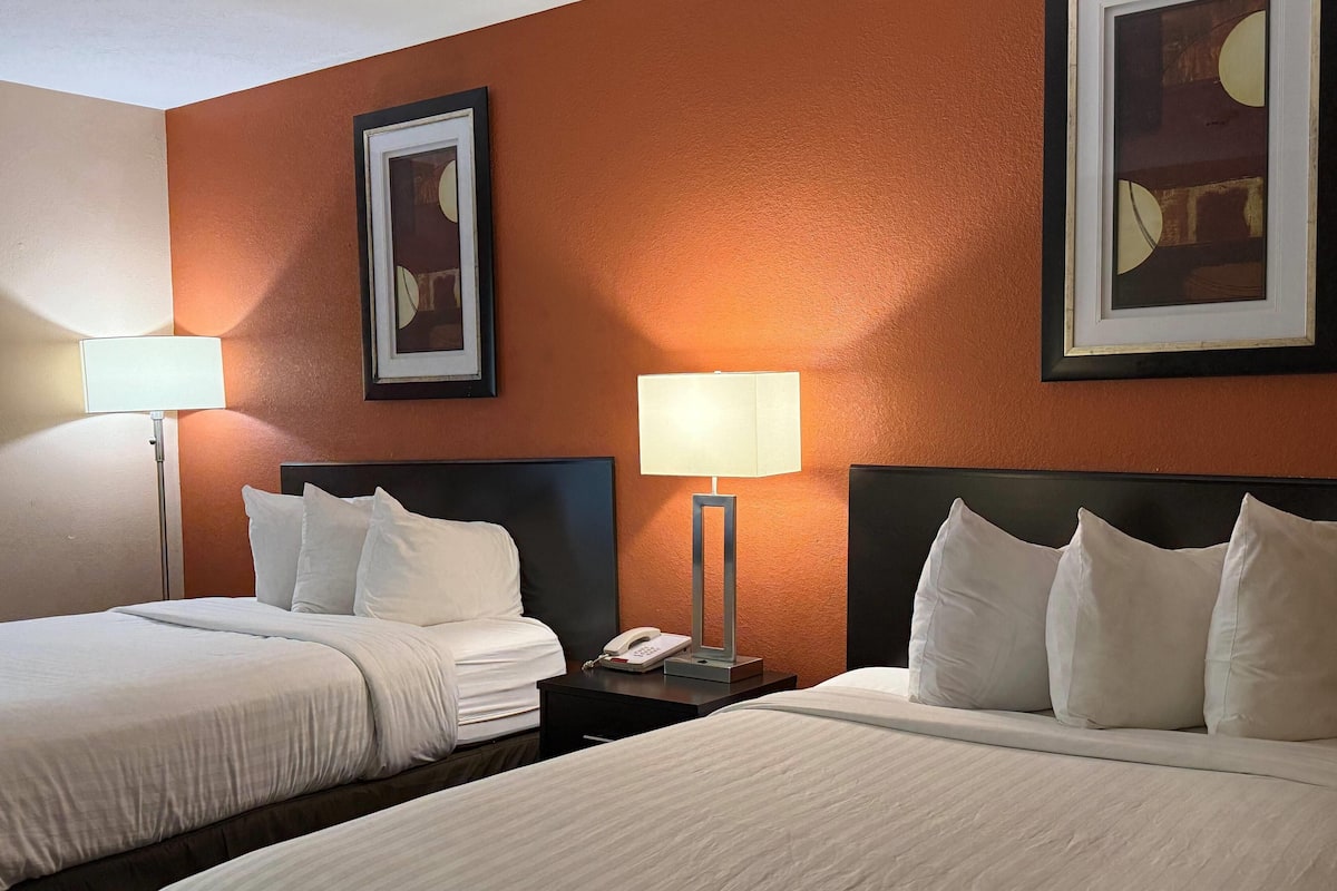 Habitaci ón de hotel con dos camas, cada una con ropa de cama y almohadas blancas, una mesita de noche con lámpara y cuadros en las paredes.