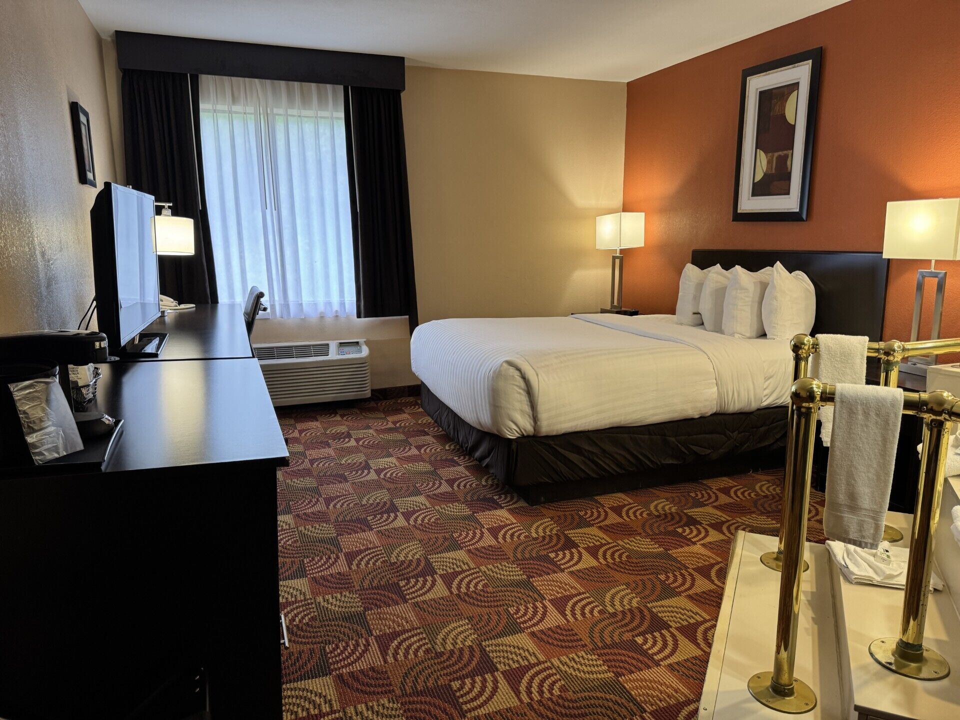 A hotel room with a bed, two bedside lamps, a desk with a computer, a TV, and a window with curtains.