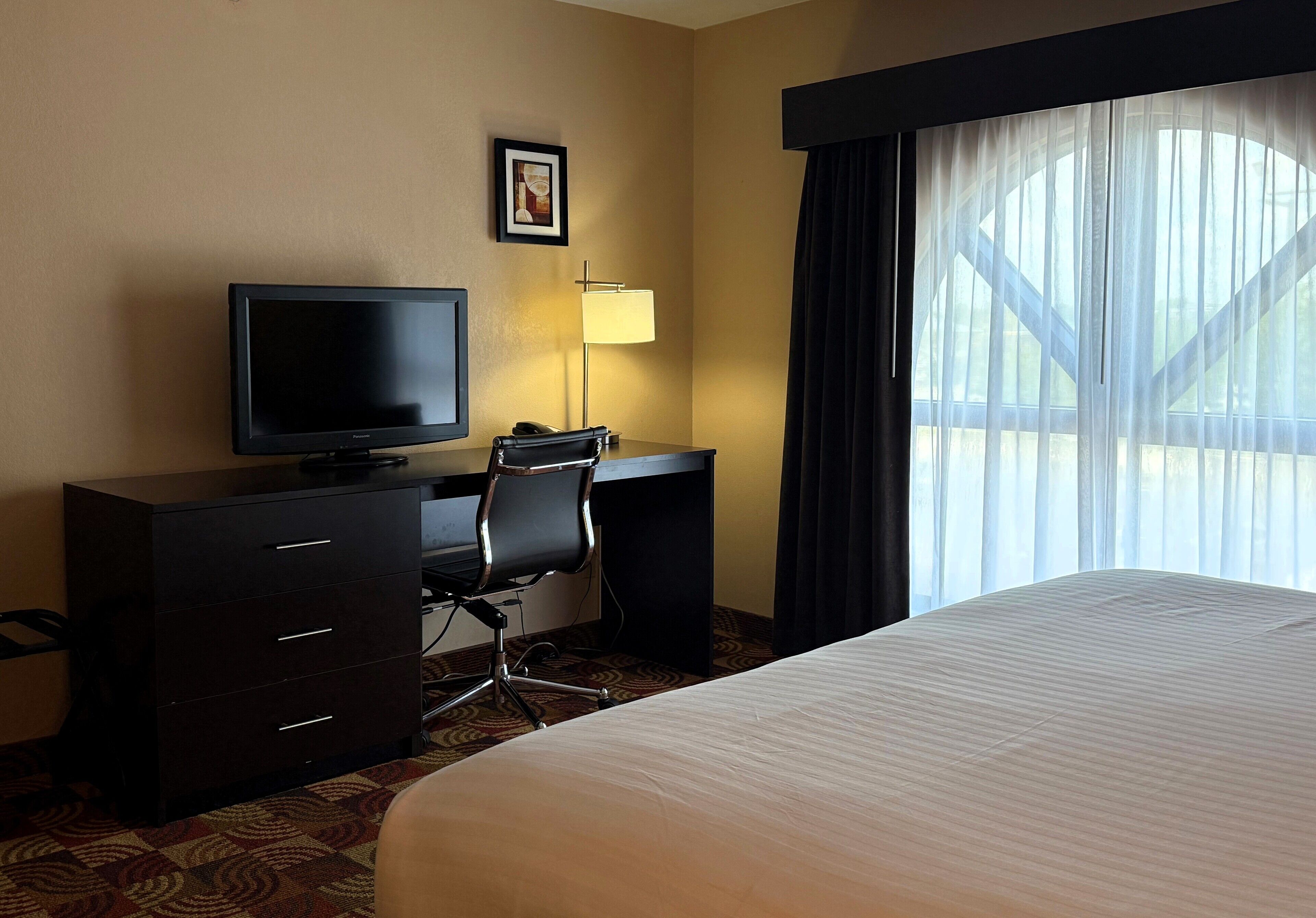 A hotel room with a bed, a desk with a computer, a chair, a television, and a window with curtains.