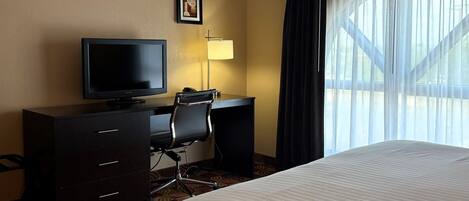 A hotel room with a bed, a desk with a computer, a chair, a television, and a window with curtains.