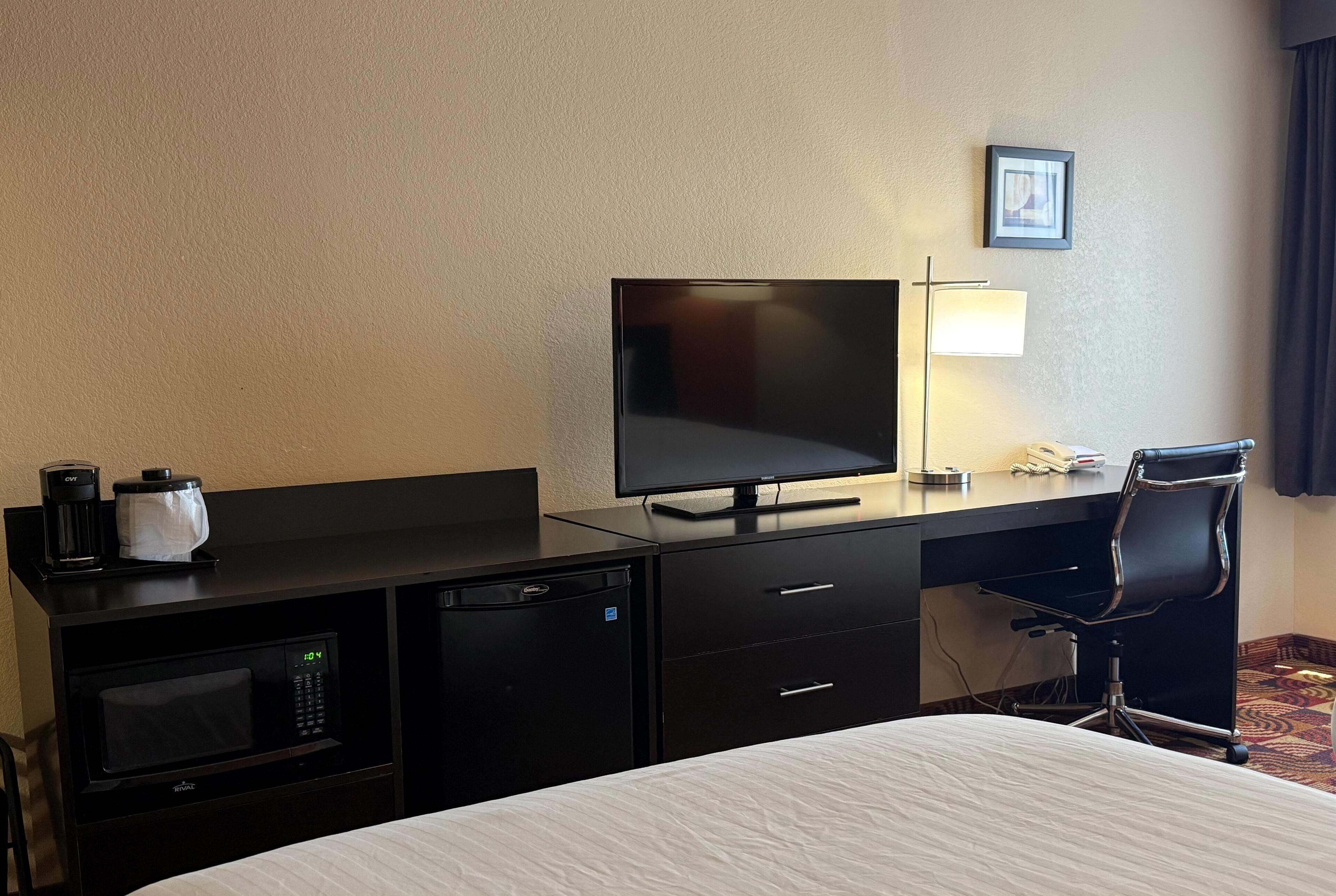 A hotel room with a bed, a desk with a flat-screen TV, a microwave, a mini-fridge, a coffee maker, a chair, and a bedside lamp.