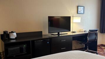 A hotel room with a bed, a desk with a flat-screen TV, a microwave, a mini-fridge, a coffee maker, a chair, and a bedside lamp.