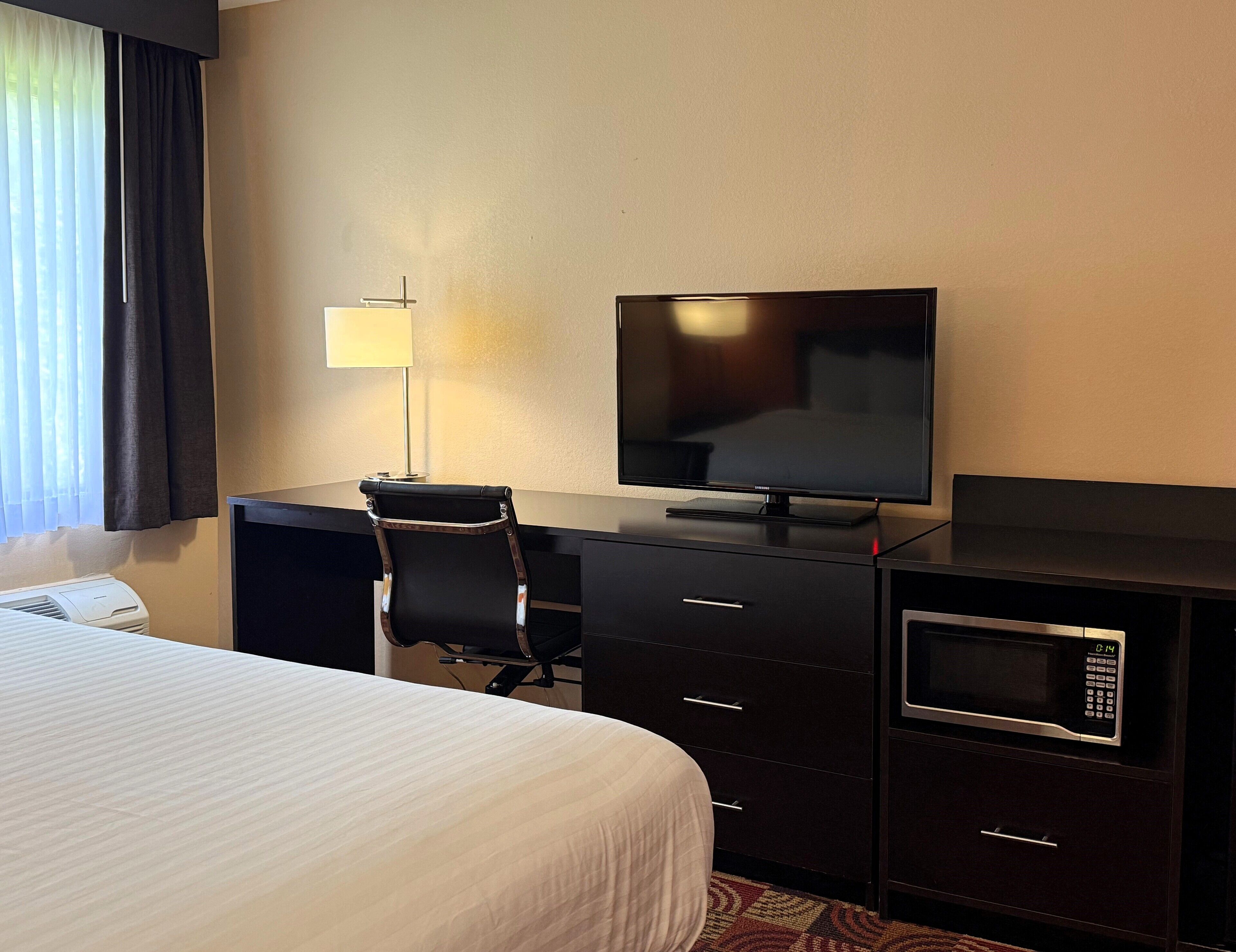 A hotel room with a bed, a desk with a flat-screen TV, a chair, a lamp, a microwave, and a dresser.