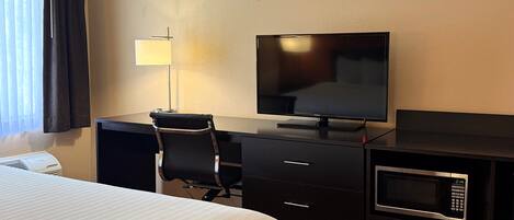 A hotel room with a bed, a desk with a flat-screen TV, a chair, a lamp, a microwave, and a dresser.