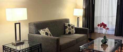 Une chambre d’hôtel avec un canapé gris, deux tables d’appoint avec des lampes, une table basse en verre et une fenêtre ornée de rideaux légers.
