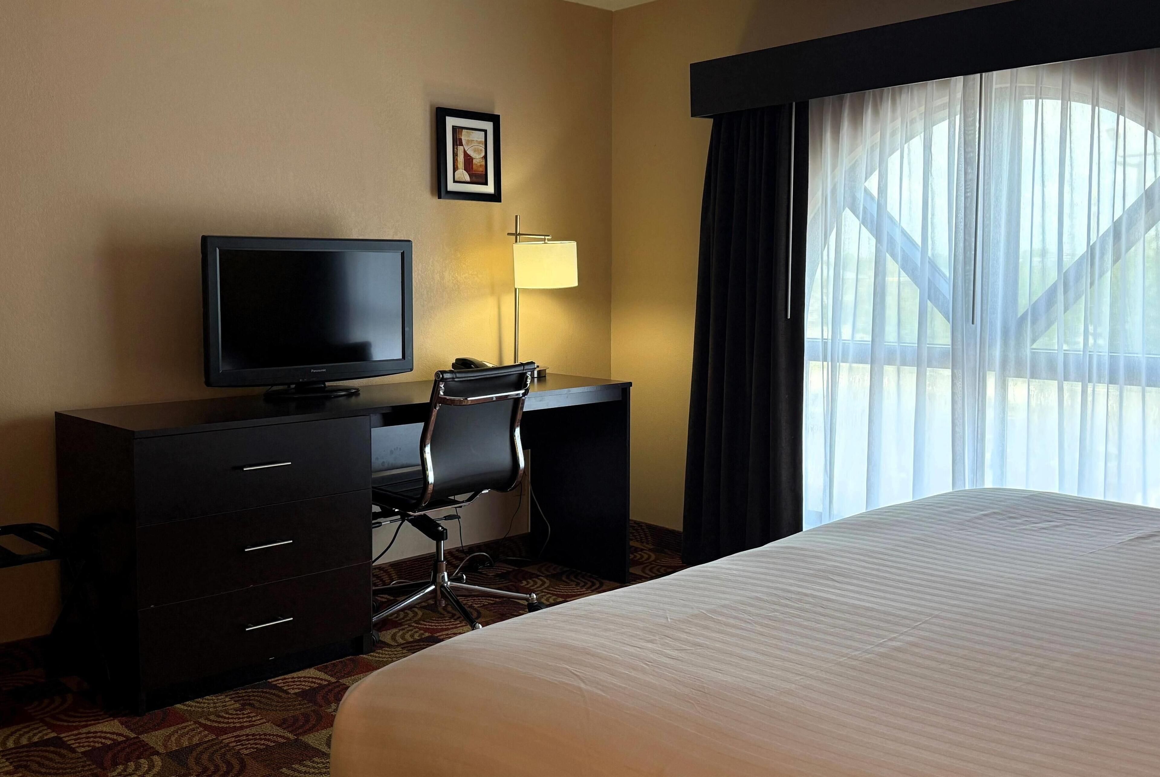 Habitación de hotel con cama, escritorio con computadora, silla, televisor y ventana con cortinas.