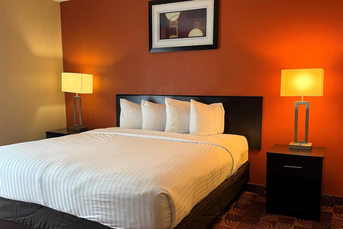 Una cama bien hecha con sábanas blancas y un cabecero oscuro, flanqueada por dos mesitas de noche con lámparas, en una habitación con paredes naranja y un cuadro enmarcado sobre la cama.