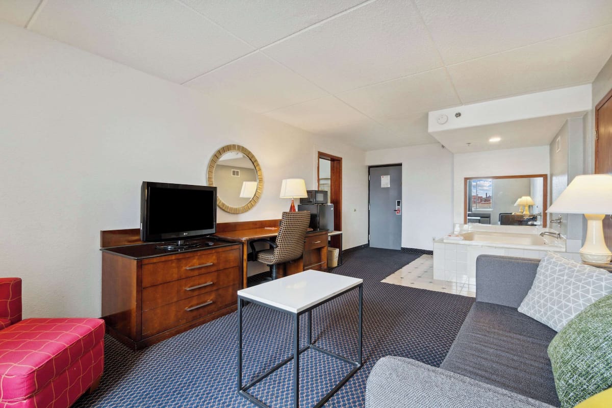 A hotel room with a TV, a desk, a chair, a sofa, a small table, a mirror, a lamp, and a bathroom with a sink and mirror.