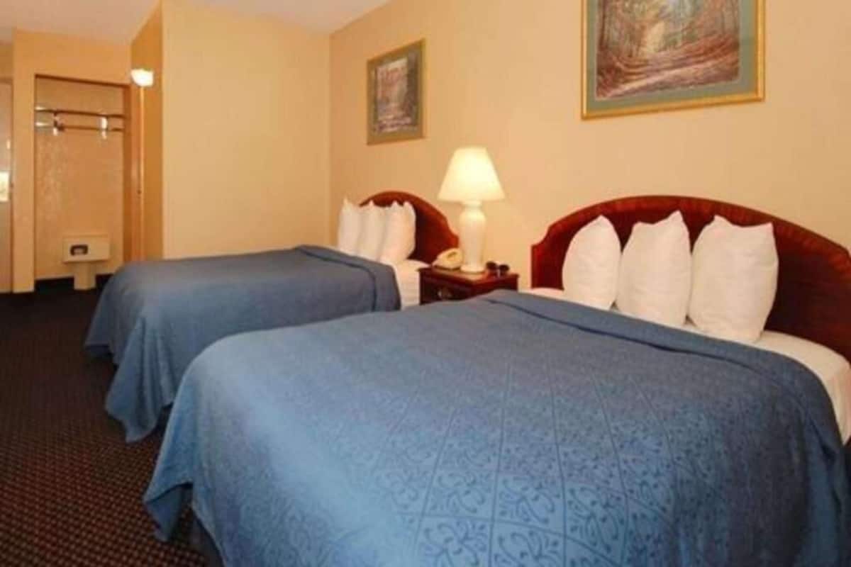 A hotel room with two beds, a nightstand with a lamp, a painting on the wall, and a door leading to a bathroom.