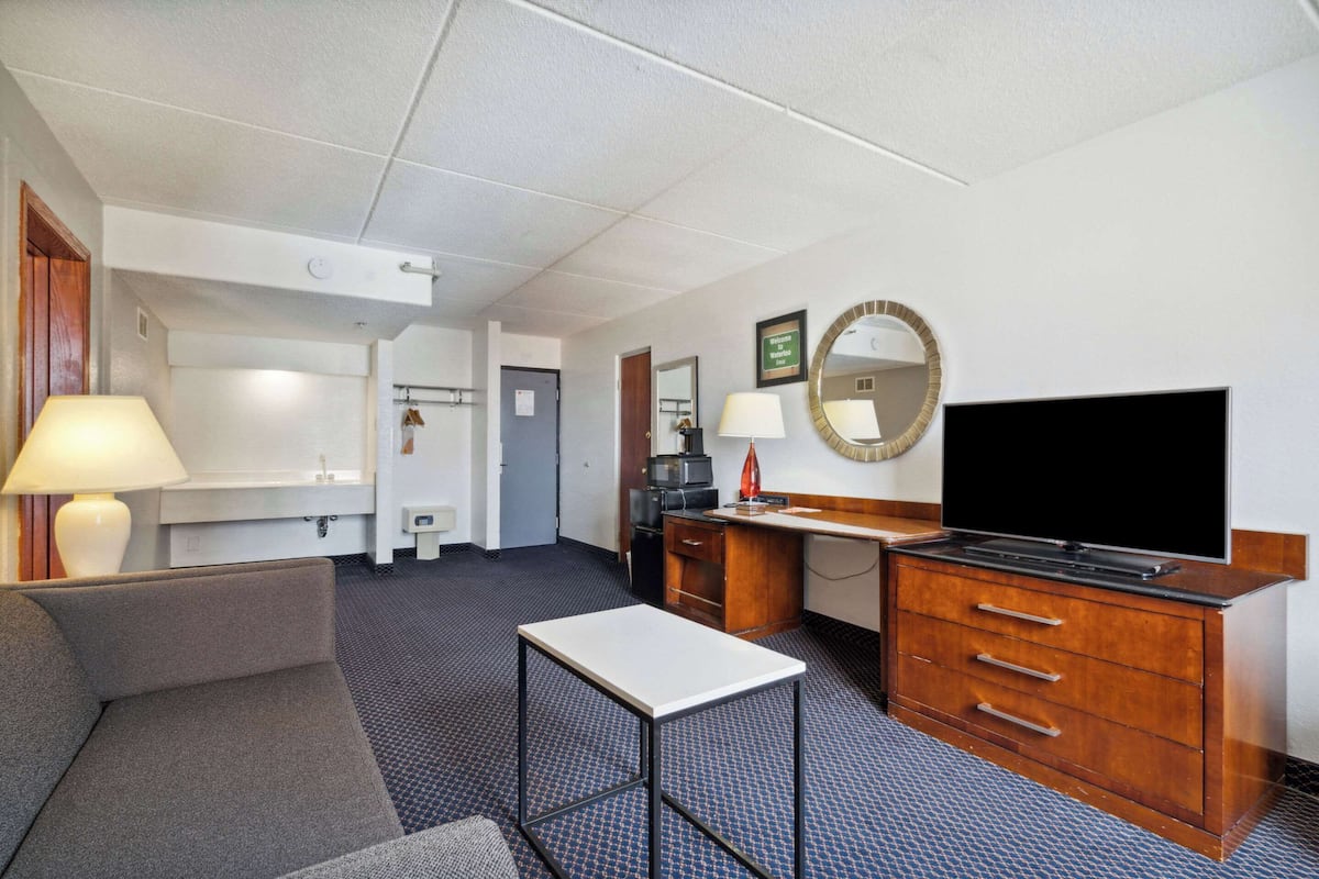 A hotel room with a sofa, a small table, a TV, a desk with drawers, a mirror, and a bathroom visible in the background.