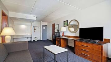 Une chambre d’hôtel avec un canapé, une petite table, une télévision, un bureau avec des tiroirs, un miroir et une salle de bain visible au fond.