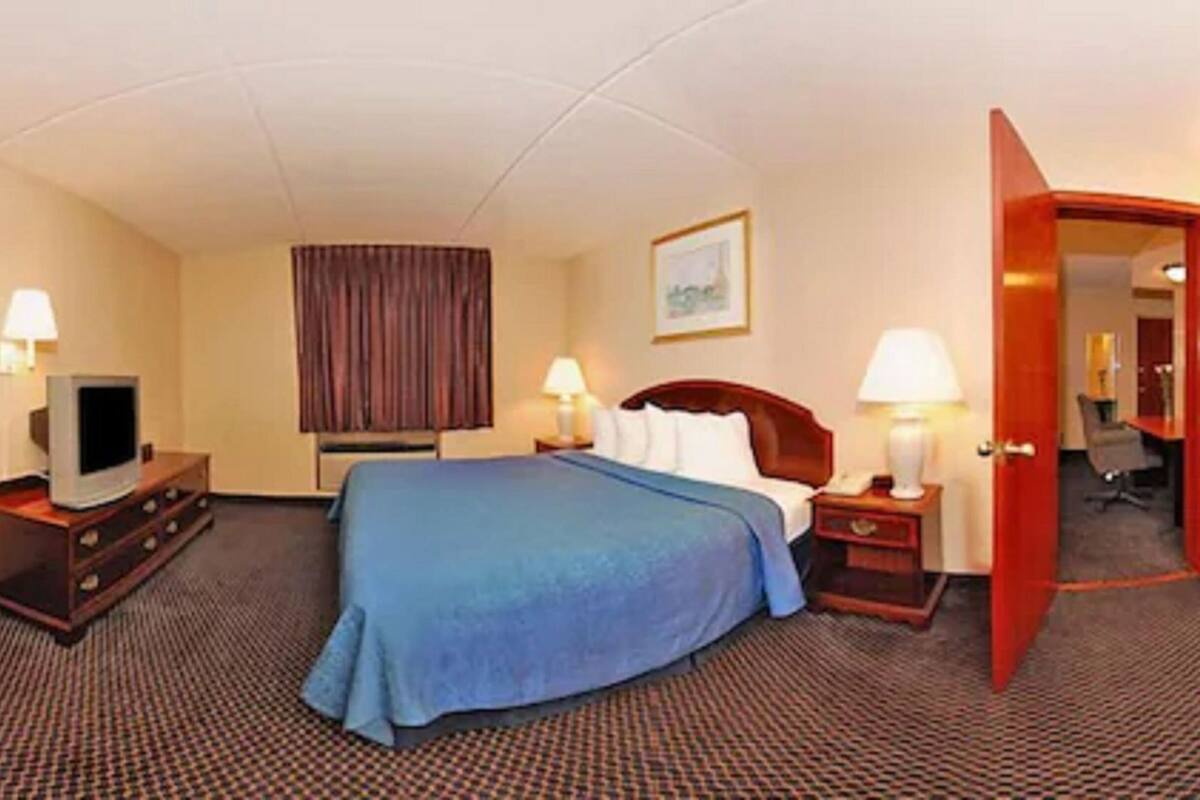 A hotel room with a bed, a TV on a dresser, two bedside tables with lamps, a door, and a carpeted floor.