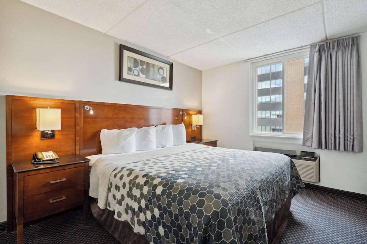 A hotel room with a bed, bedside lamps, a framed picture on the wall, a window with curtains, and a thermostat on the wall.