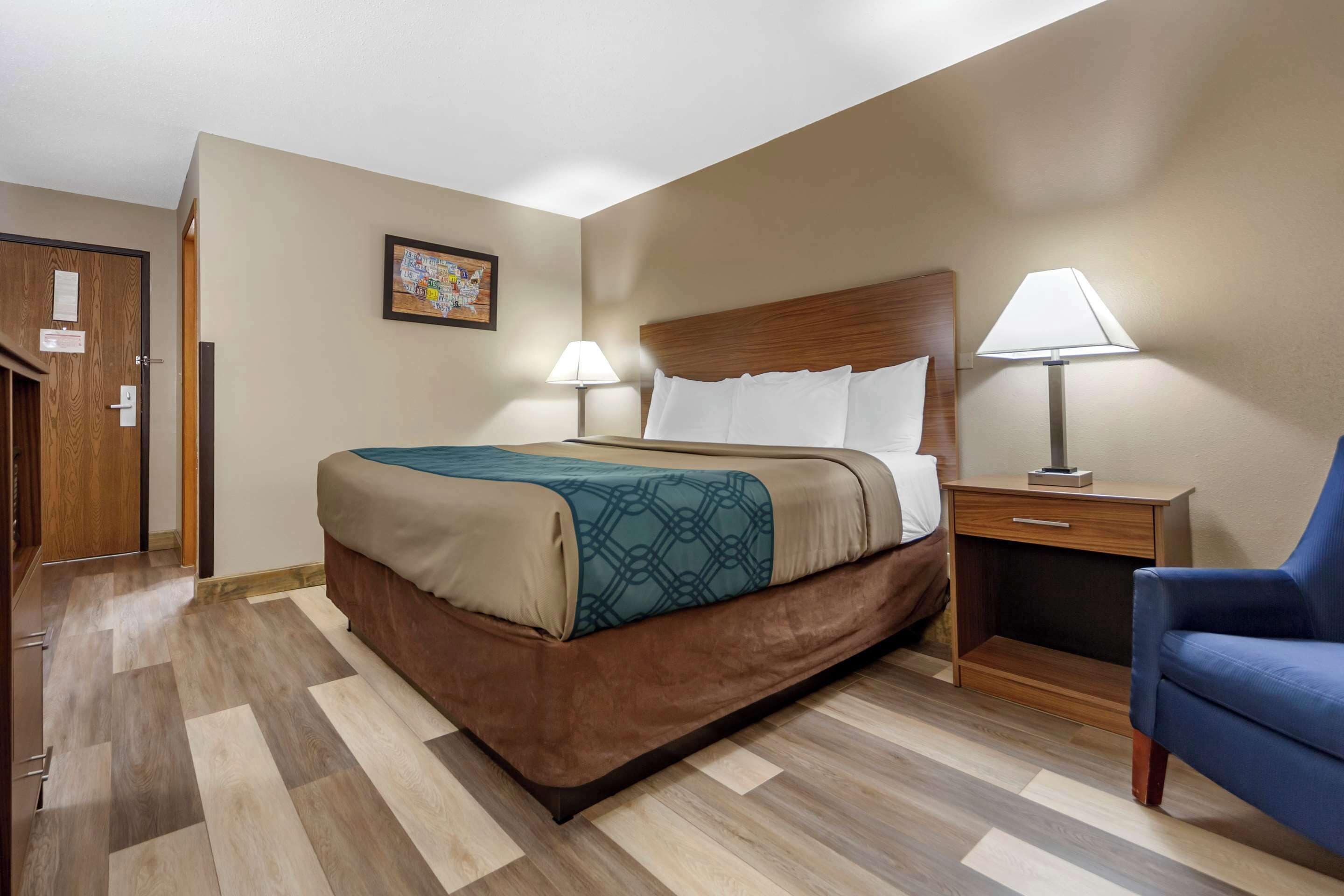 A hotel room with a large bed, two bedside tables with lamps, a wooden headboard, and a blue armchair.