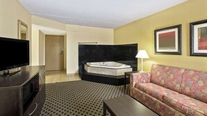 A hotel room with a large flat-screen TV, a sofa, a coffee table, a patterned rug, a black bathtub, and framed artwork on the wall.