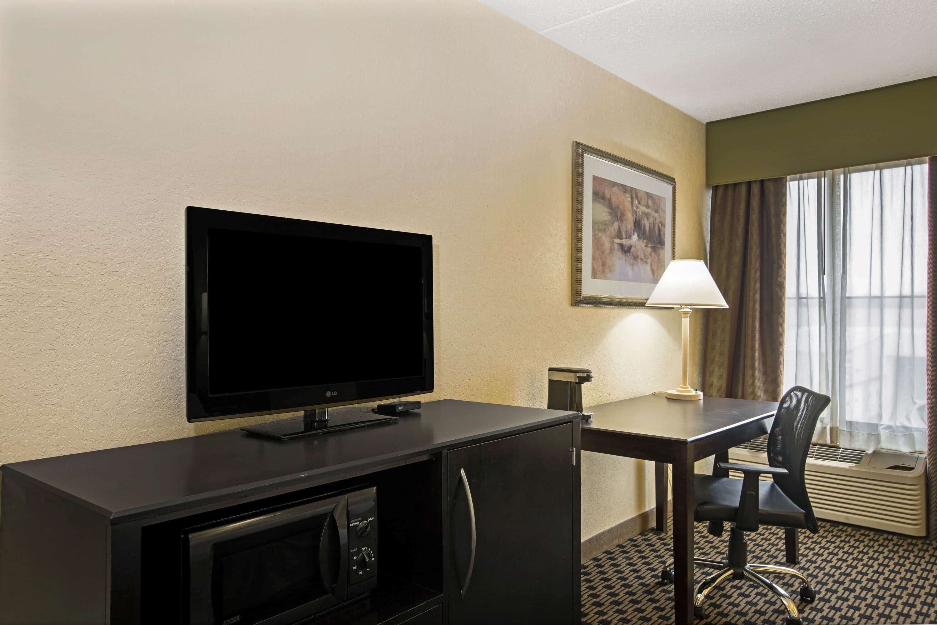 A hotel room with a flat-screen TV, a desk with a chair, a lamp, and a framed picture on the wall.