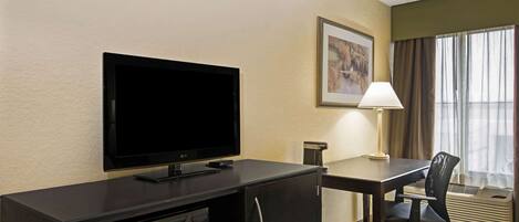 A hotel room with a flat-screen TV, a desk with a chair, a lamp, and a framed picture on the wall.
