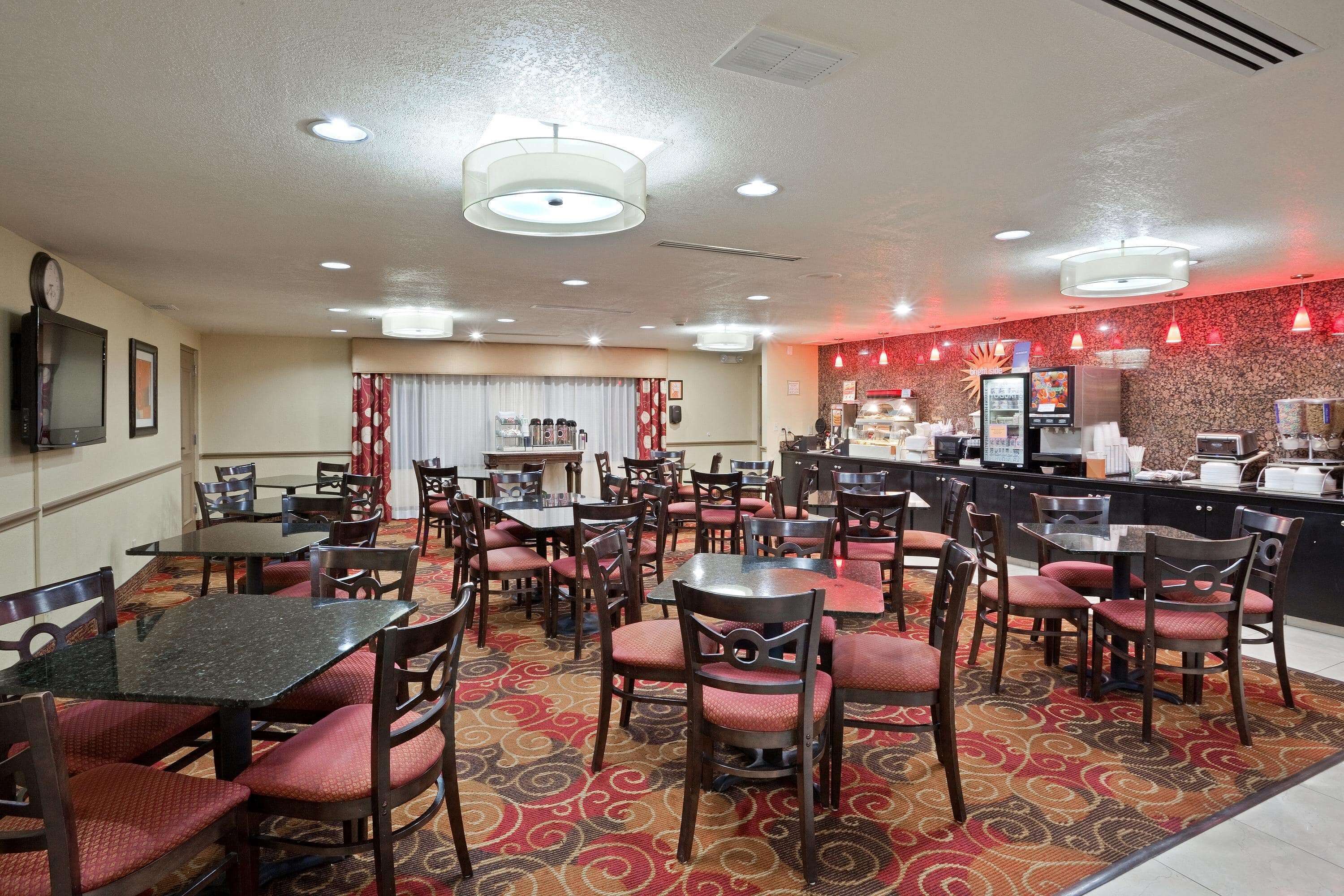 A hotel dining area with multiple tables and chairs, a kitchen area with a counter and appliances, and a patterned carpet.