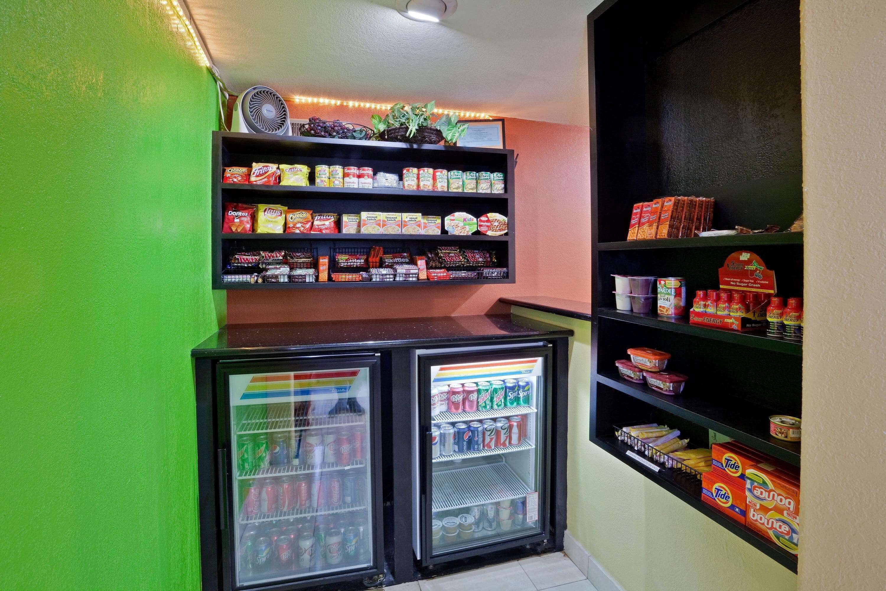 Quarto pequeno com parede verde, geladeira repleta de bebidas e prateleiras com lanches e temperos.