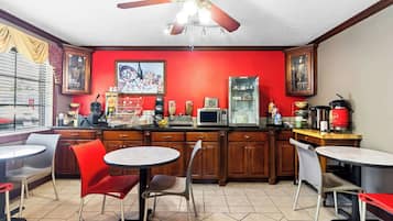 Una cocina con paredes rojas, armarios de madera y un ventilador de techo. Hay dos mesas redondas y varias sillas.