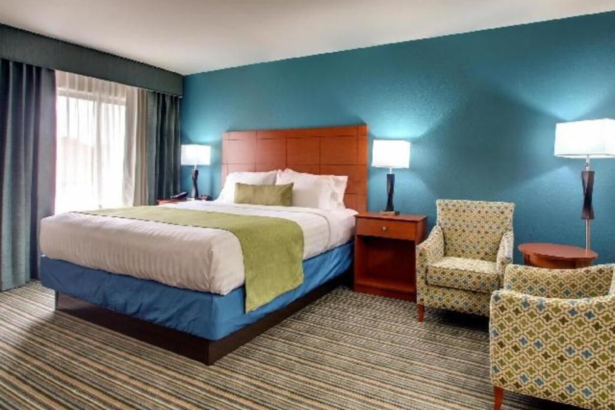 Una habitación de hotel con una cama, dos mesitas de noche, una silla y un ventanal con cortinas.