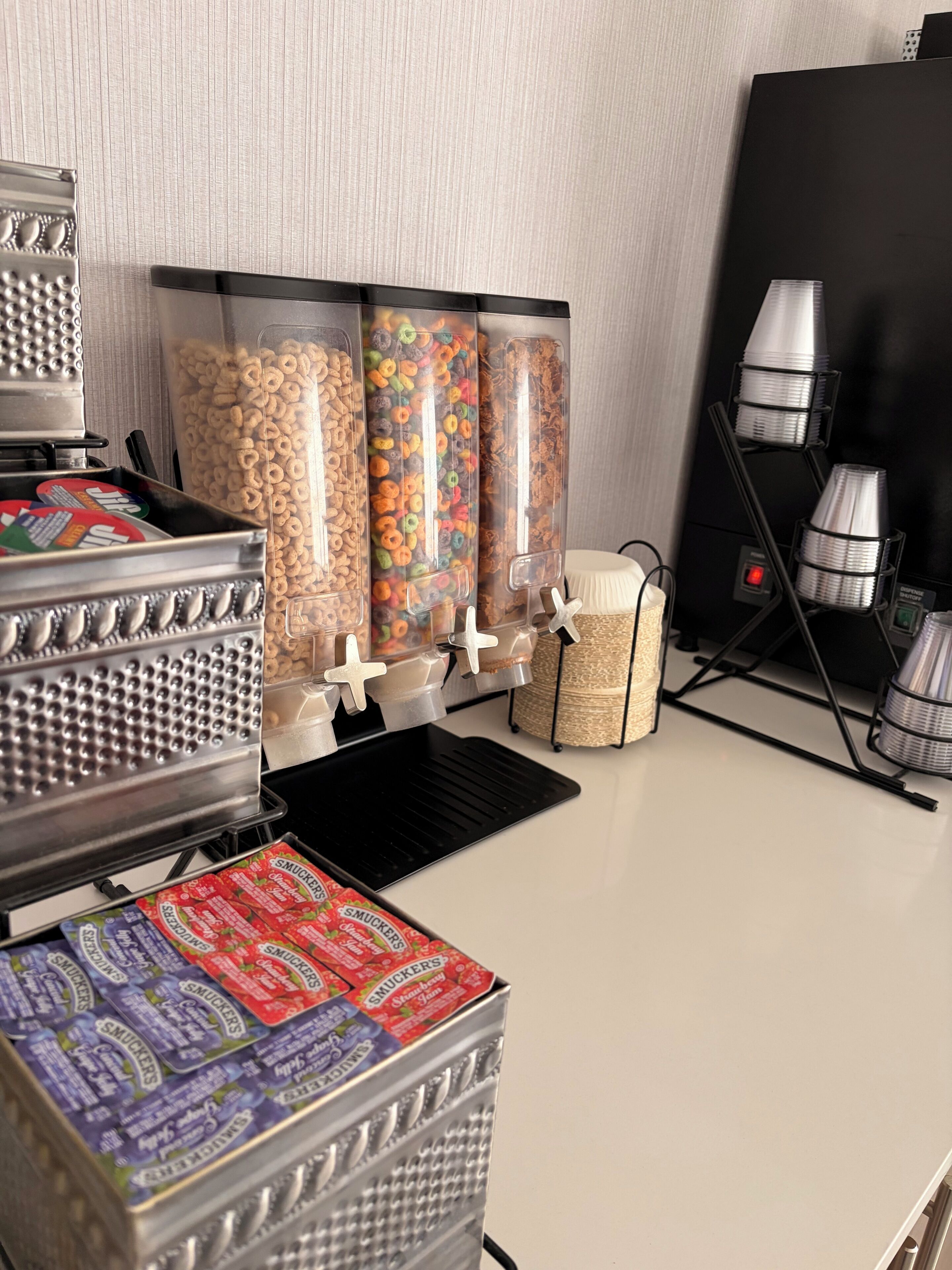 Un mostrador de aperitivos con dispensadores de cereales y dulces, una máquina para gofres y una caja de caramelos de la marca Smucker's.
