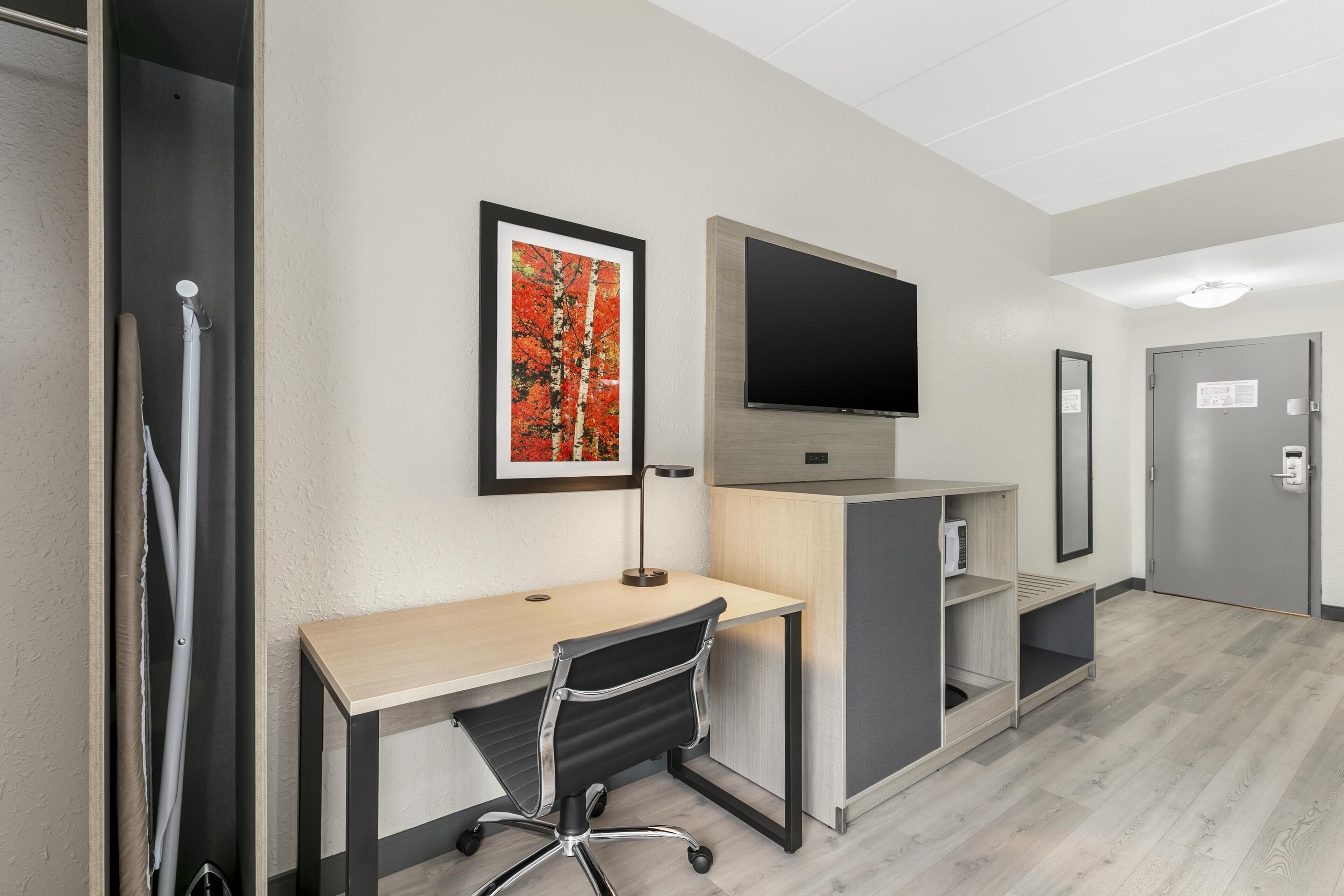 A hotel room with a desk, chair, TV, artwork, and a door.