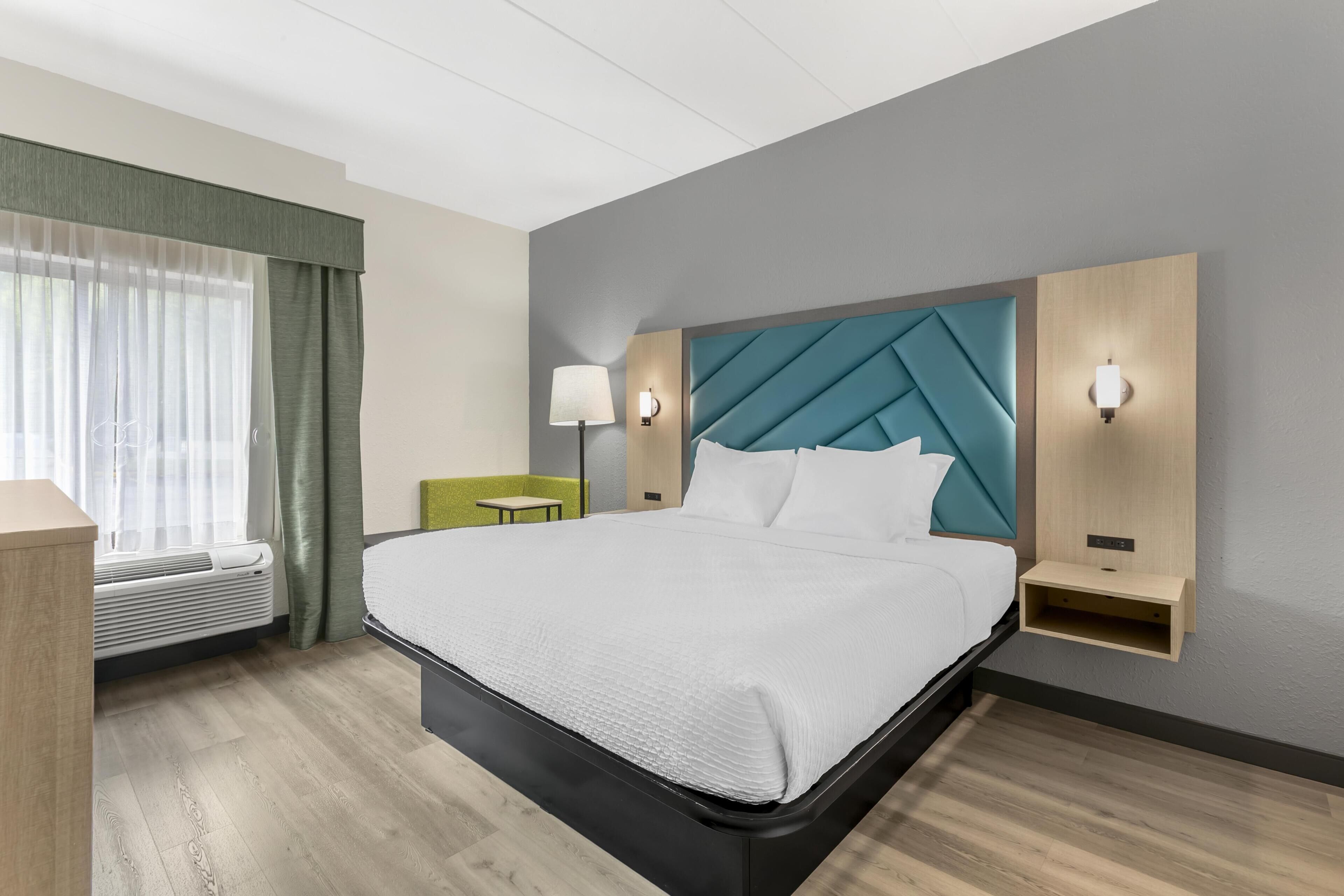 Hotellihuone, jossa on sänky, työpöytä, lamppu ja seinälle kiinnitetty hylly.