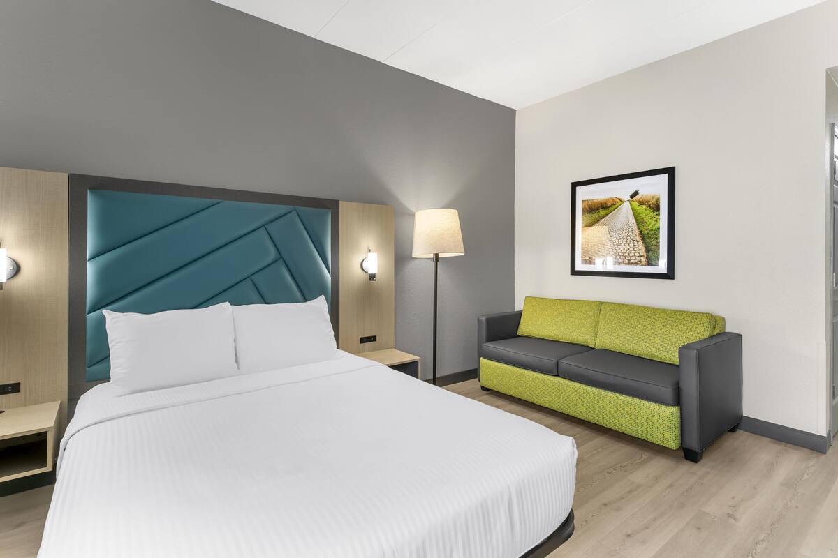 A modern hotel room with a large bed, a green and gray sofa, a wall-mounted picture, and a floor lamp.